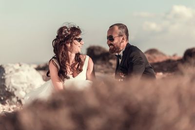 Smiling couple wearing sunglasses talking on field against sky during sunny day