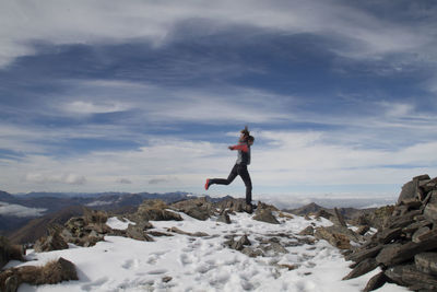 Young woman jumping on cliff against cloudy sky during winter