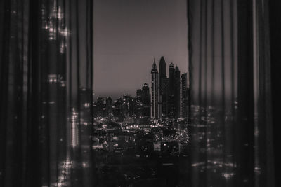 Illuminated buildings in city against sky seen through glass window