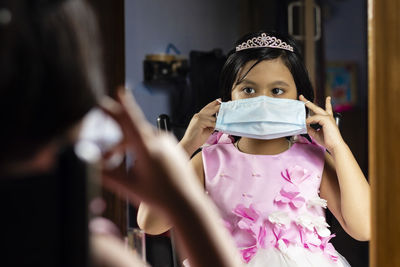 A cute indian girl child in red dress holding surgical nose mask in front of mirror