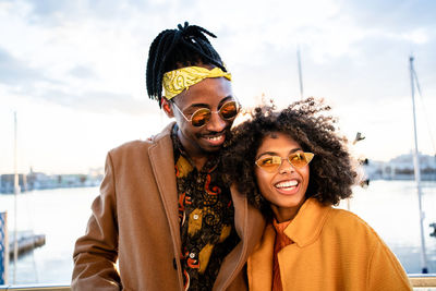 Bright laughing black woman in yellow coat with trendy boyfriend in sunglasses standing closely together looking away on urban pier