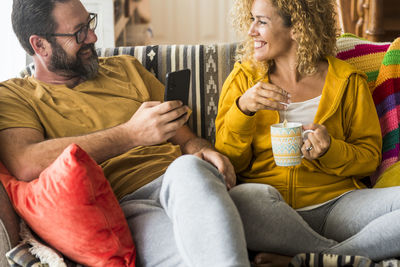Smiling man holding mobile phone sitting by woman with cup on sofa