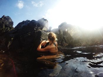 Young woman in bikini sitting on rock against sky