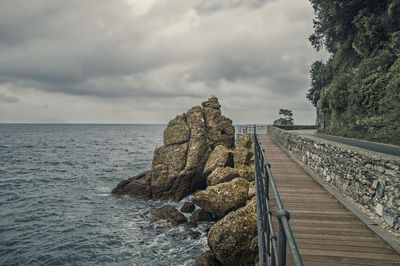 Narrow footbridge along calm sea against cloudy sky