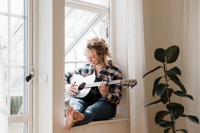 Woman sat at home on a window ledge smiling playing guitar