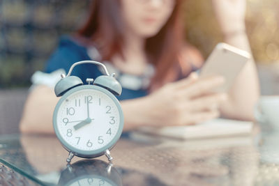 Close-up of alarm clock with woman using phone on table