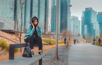 Full length portrait of young woman sitting against modern building in city
