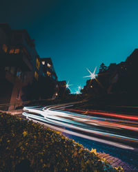 Light trails on road by buildings against sky at night