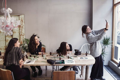 Smiling owner taking selfie with female coworkers sitting at table in perfume workshop