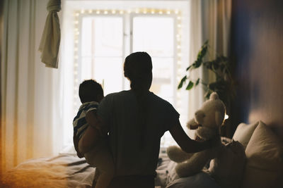 Woman keeping teddy bear on bed while carrying son at bedroom