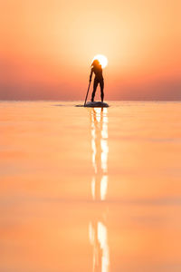 Back view of silhouette of unrecognizable female surfer standing on paddleboard and rowing against spectacular sun in sunset sky