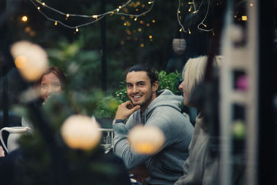 Smiling young man sitting amidst friends at dining table in back yard during garden party