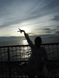 Woman with arms raised standing at sea shore against sky during sunset