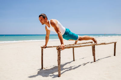 Man exercising on parallel bars at beach against blue sky