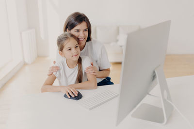 Smiling woman with daughter using desktop pc at table