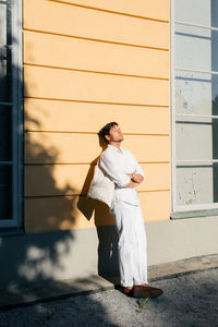 Full length of young man looking away, enjoying the sunlight while standing against building