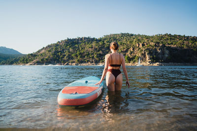 Back view of unrecognizable female in swimwear standing near surfboard in lake against forest on sunny summer day