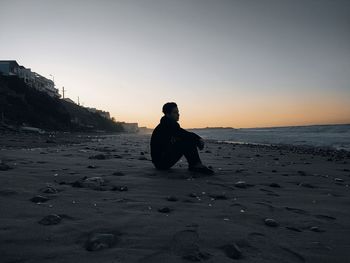 Man sitting on beach against sky during sunset