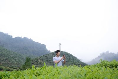 Man photographing with mobile phone while standing amidst tea crops on field against clear sky