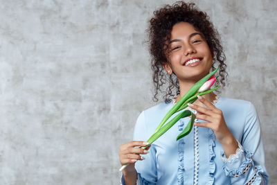 Portrait of smiling young woman holding tulip while standing against wall