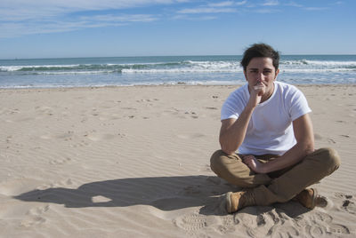 Young man sitting at beach against sky