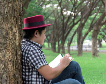 Side view of man writing in diary against tree trunk at public park