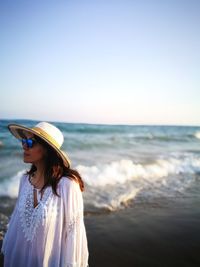 Young woman wearing hat and sunglasses while standing against sea and sky