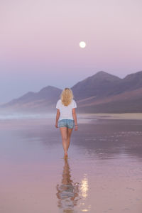 Rear view of man standing on beach against sky during sunset
