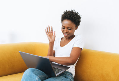 Beautiful young african american woman in white t-shirt and blue jeans using laptop on yellow sofa 