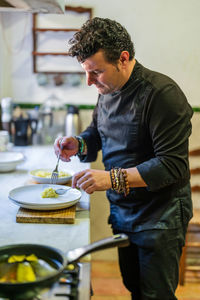 Side view of concentrated male cook in black uniform with bracelets serving tasty food on plate in modern kitchen of restaurant