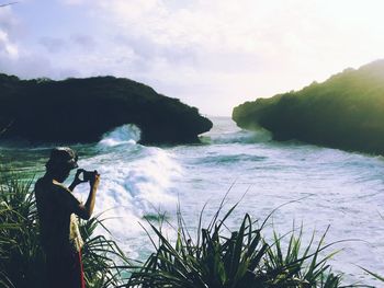 Man photographing woman standing by sea against sky