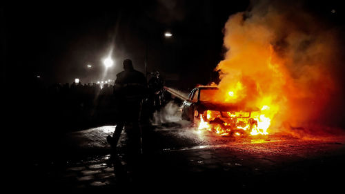 Firefighters standing by burning car on street at night