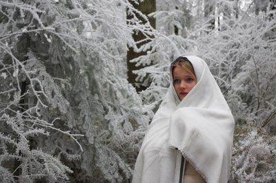 Woman wrapped in blanket looking away while standing by snow covered trees