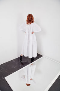 Back side view woman standing against white wall, full length fashion image  model in oversize dress