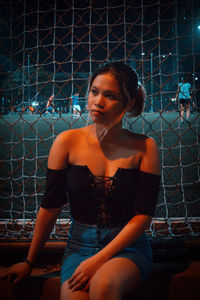 Young woman looking away while sitting on chainlink fence