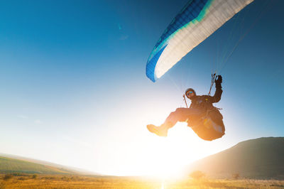 A paraglider takes off from a mountainside with a blue and white canopy and the sun behind. a