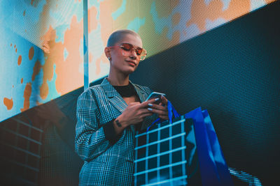 Low angle view of lesbian woman using smart phone holding shopping bags standing against abstract backgrounds