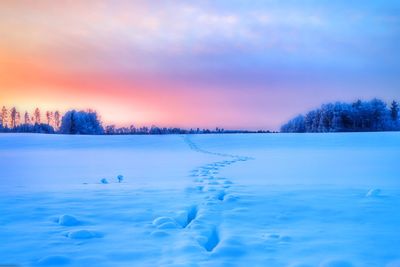 Footprints on snow covered field against sky during sunset