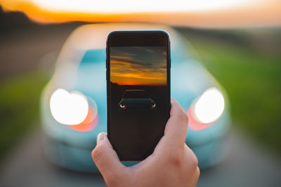 Close-up of hand photographing car against sky during sunset
