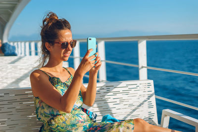 Young woman using mobile phone while sitting on railing against sea