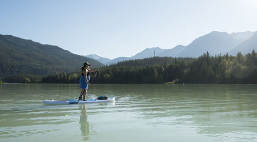 Barefoot woman riding paddle board on lake against green mountain ridge on sunny summer day in british columbia, canada