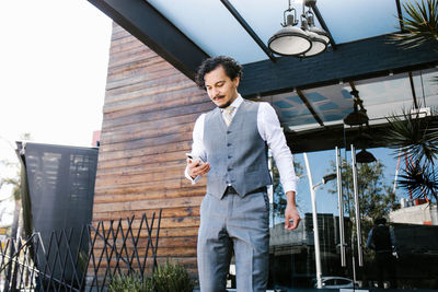 Serious bearded male entrepreneur in formal suit browsing cellphone while standing on veranda of modern building on street in city