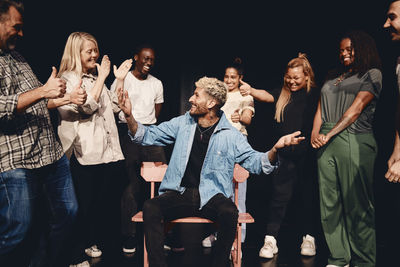 Happy young man sitting on chair amidst multiracial stage performers in class