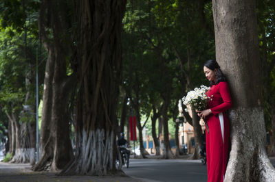 Woman holding flower bouquet while standing against tree trunk