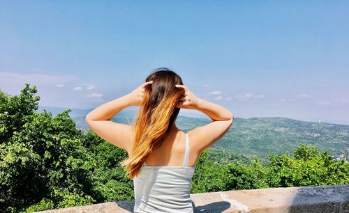 Rear view of young woman with hand in hair standing at observation point against blue sky during sunny day