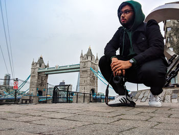 Full length of male tourist holding camera while crouching on footpath with tower bridge in background
