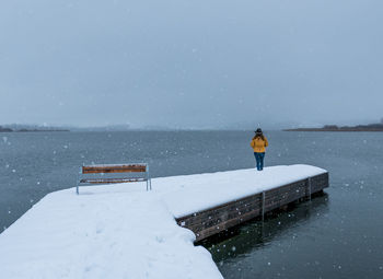 Rear view of woman standing at lake against sky during snowfall