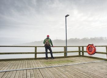 Rear view of man standing on pier over lake against sky