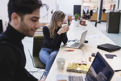 Young woman drinking coffee while sitting with colleague at desk in office