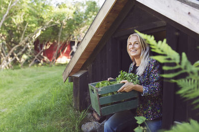 Smiling woman carrying vegetables crate while coming out of cottage